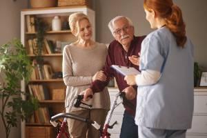 An elderly man using a walker and his wife speak with an in-home skilled nurse.