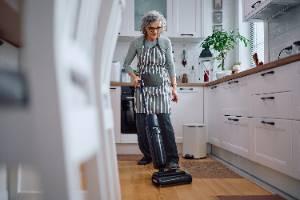 An image of an older woman vacuuming in her kitchen.
