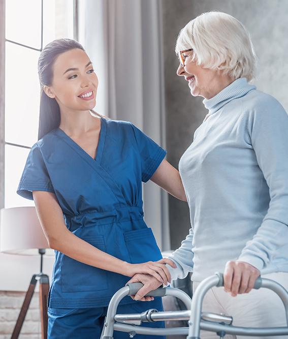 Young female social worker helping senior woman to walk with walker at home