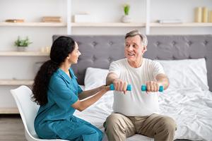 Young rehabilitation therapist assisting senior male patient with strength exercises with dumbbells on bed at home