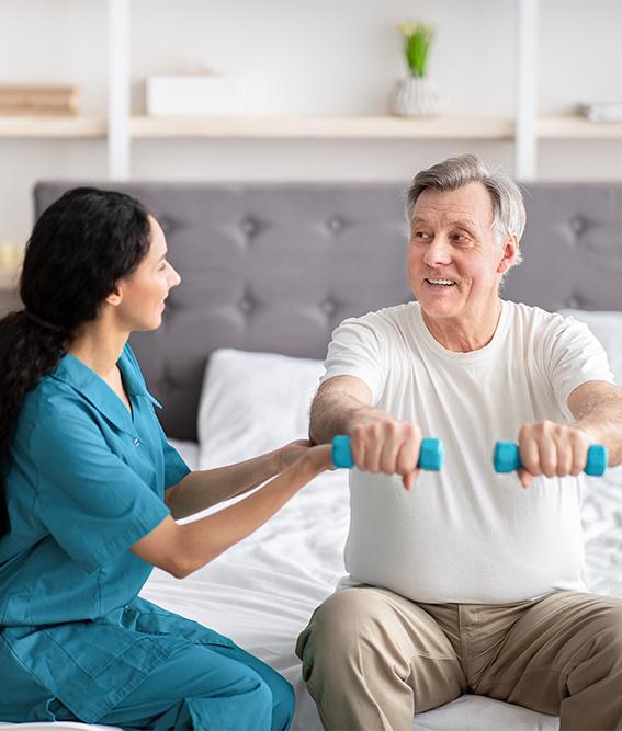 Young rehabilitation therapist assisting senior male patient with strength exercises with dumbbells on bed at home
